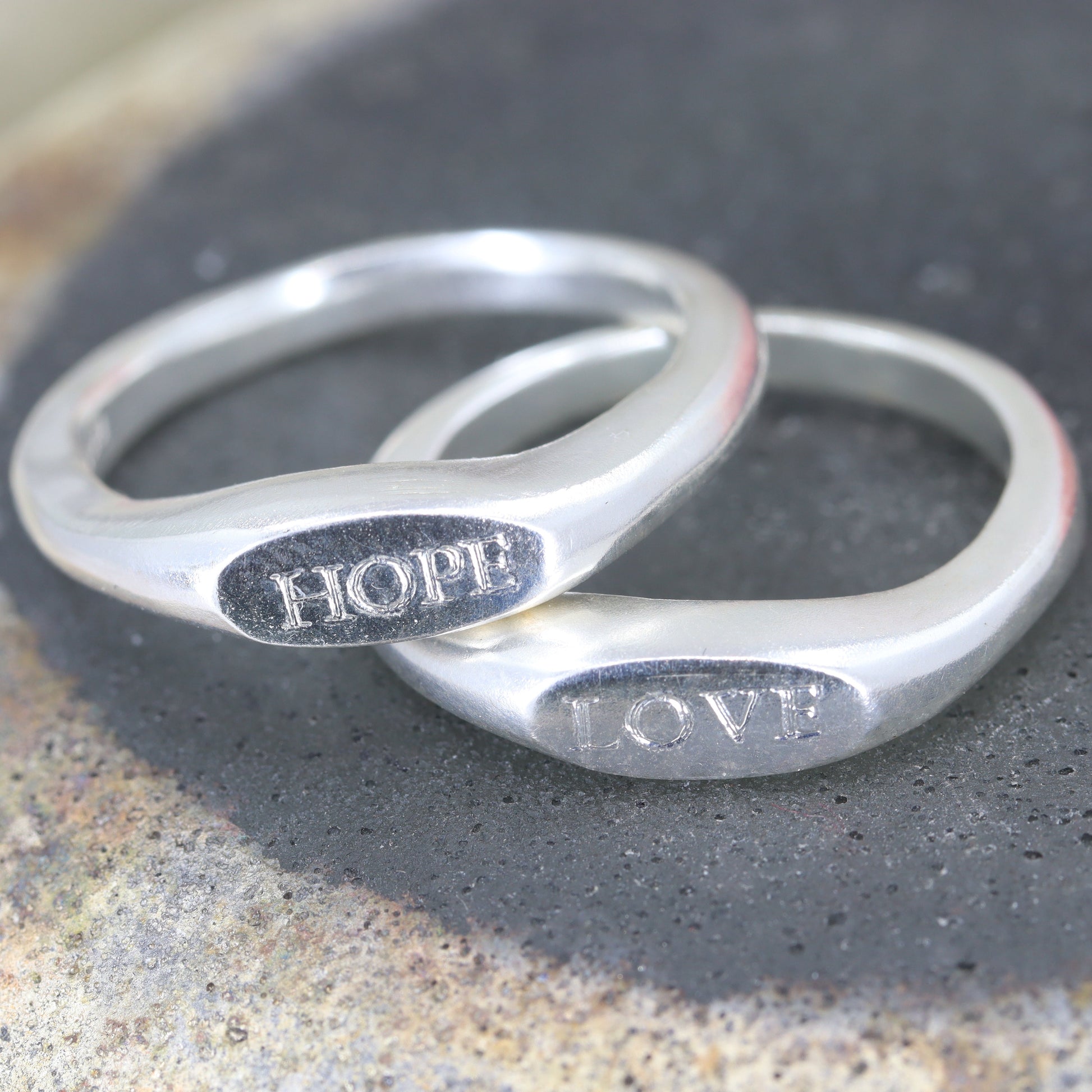Two silver rings with 'HOPE' and 'LOVE' engraved on them, lying on a textured surface.